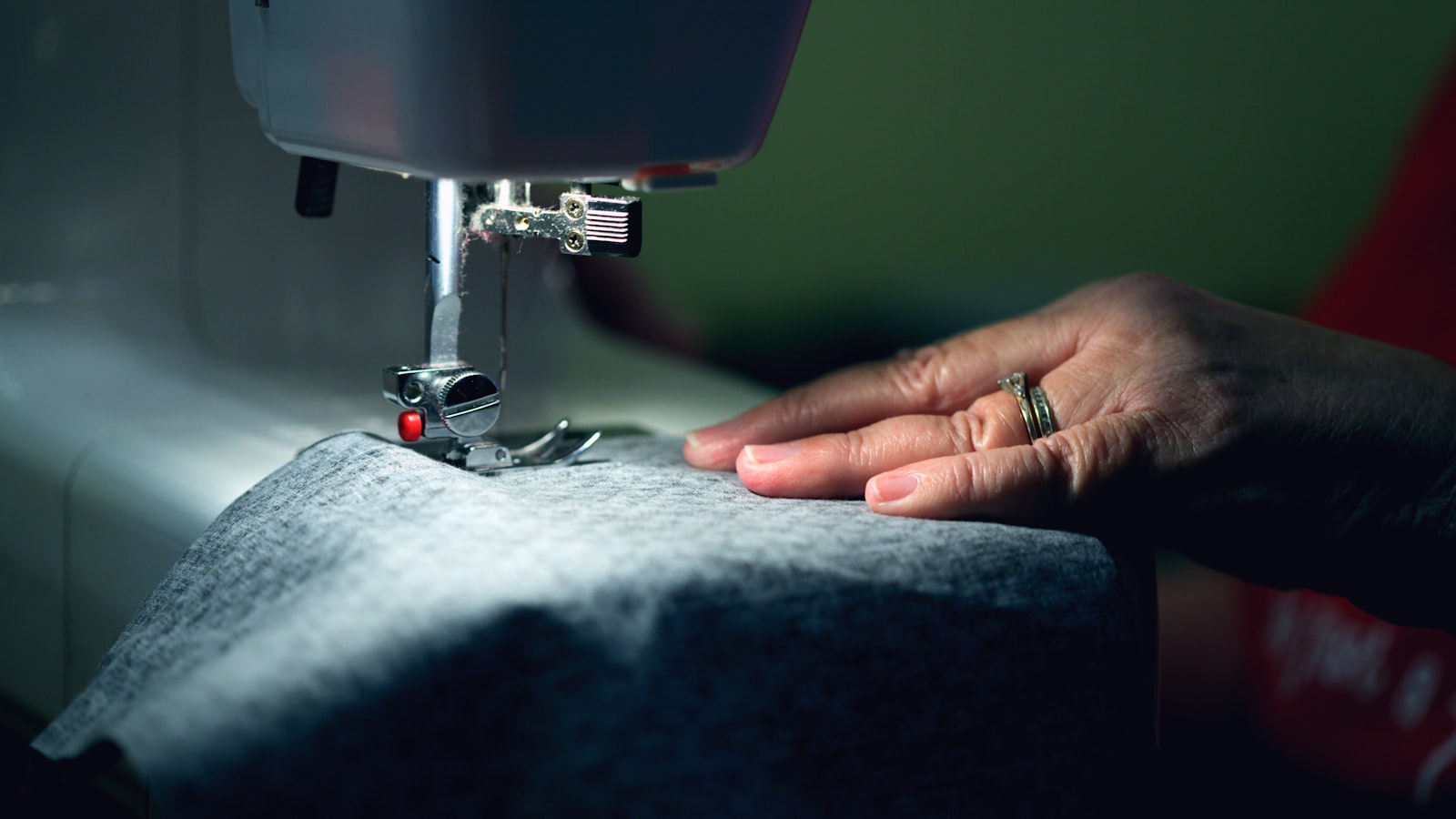 A hand with rings guides gray fabric under the needle of a lit sewing machine
