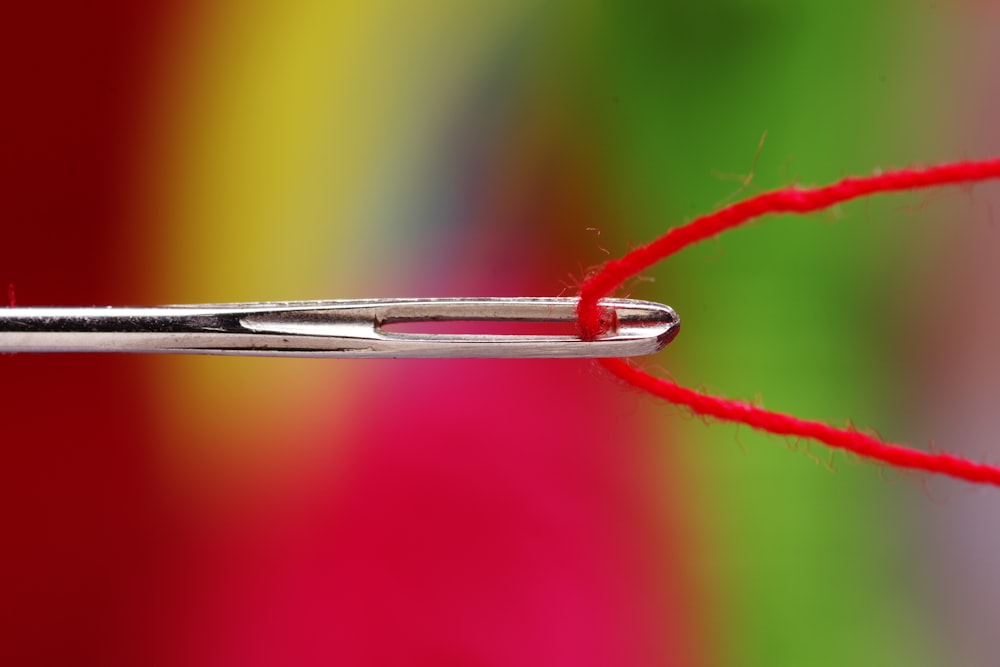 A close-up of a silver sewing needle threaded with red yarn, against a blurry multicolored background