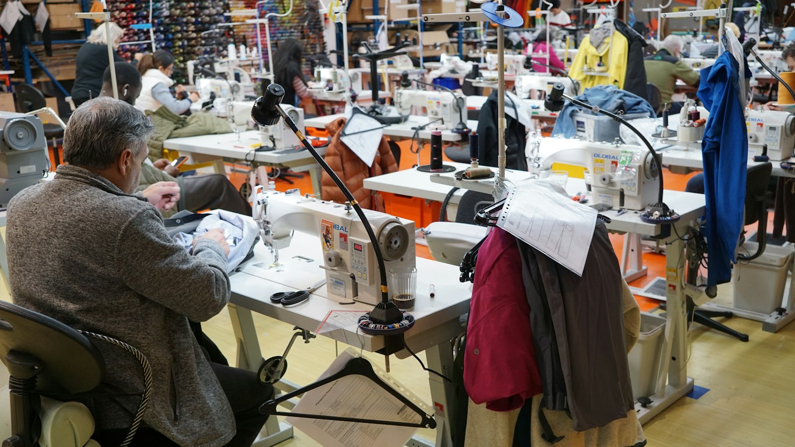 A man in a grey sweater sits at a sewing machine in a large workshop with other workers