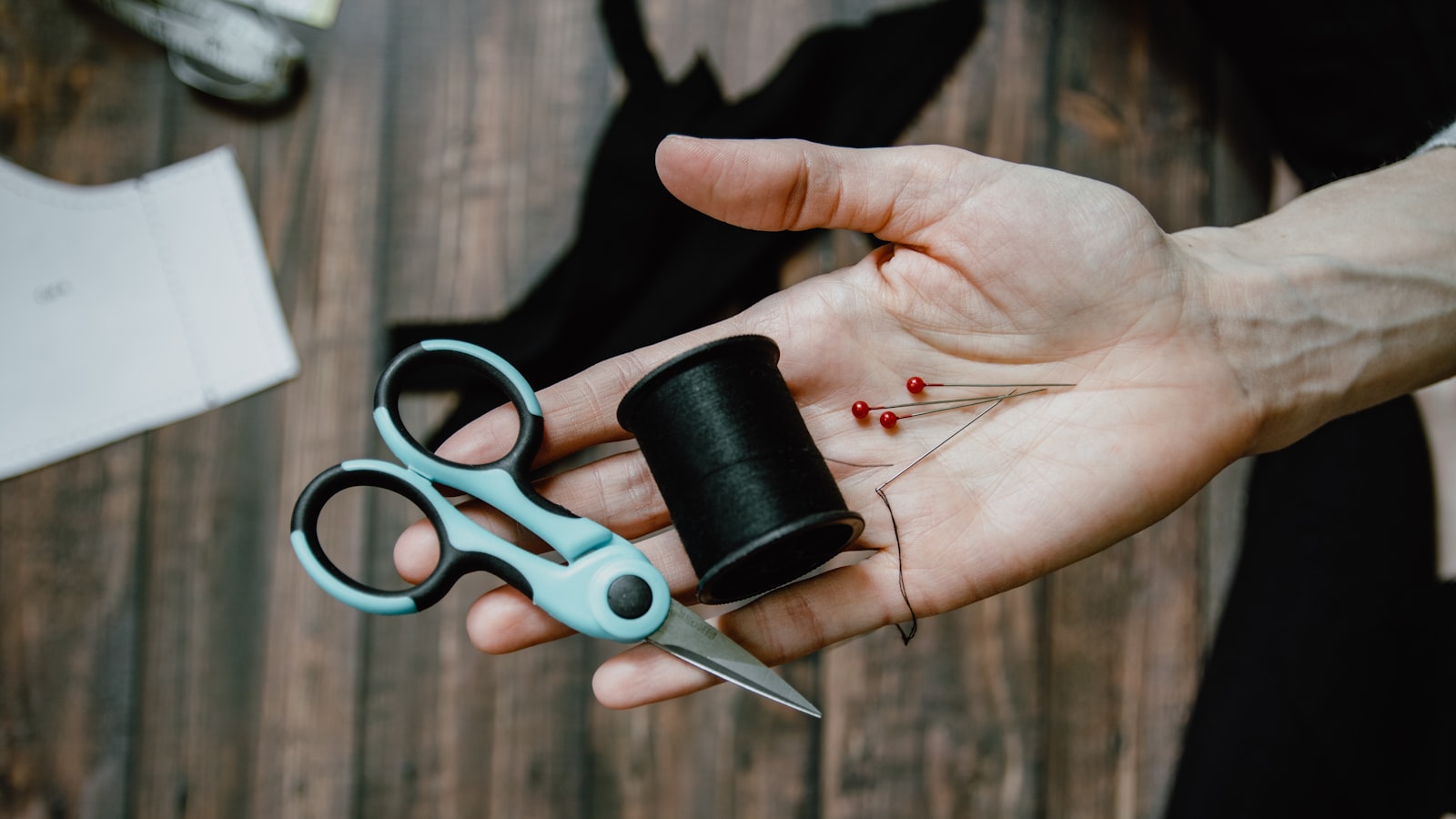 A hand holds small scissors, a black spool of thread, pins, and a needle above a wooden table
