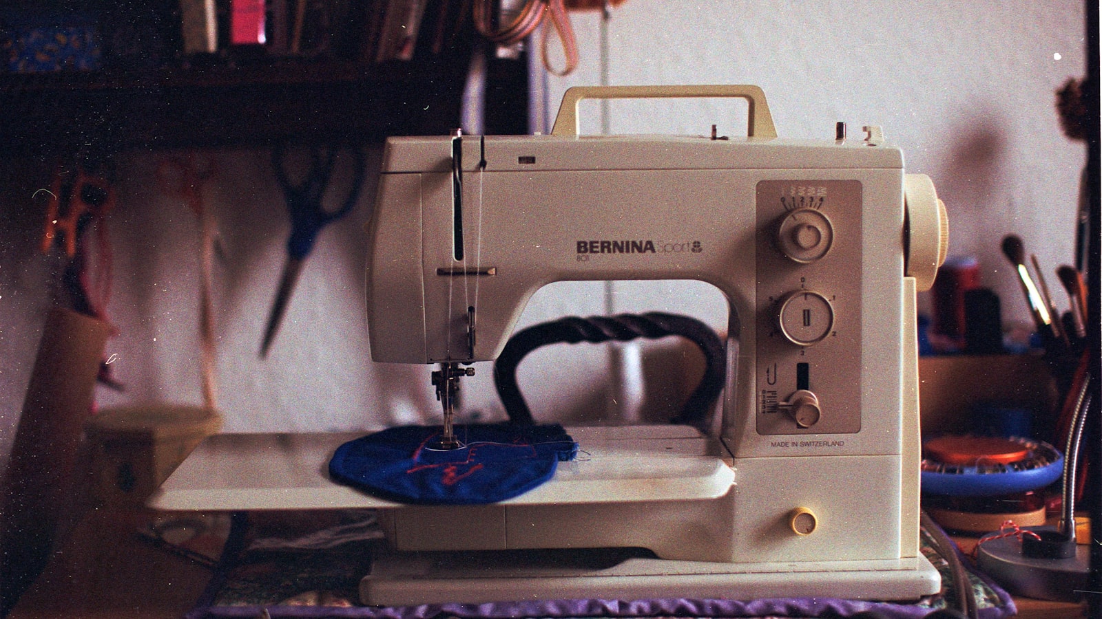 A Bernina sewing machine sits on a table, threaded with white thread and stitching a blue piece of fabric
