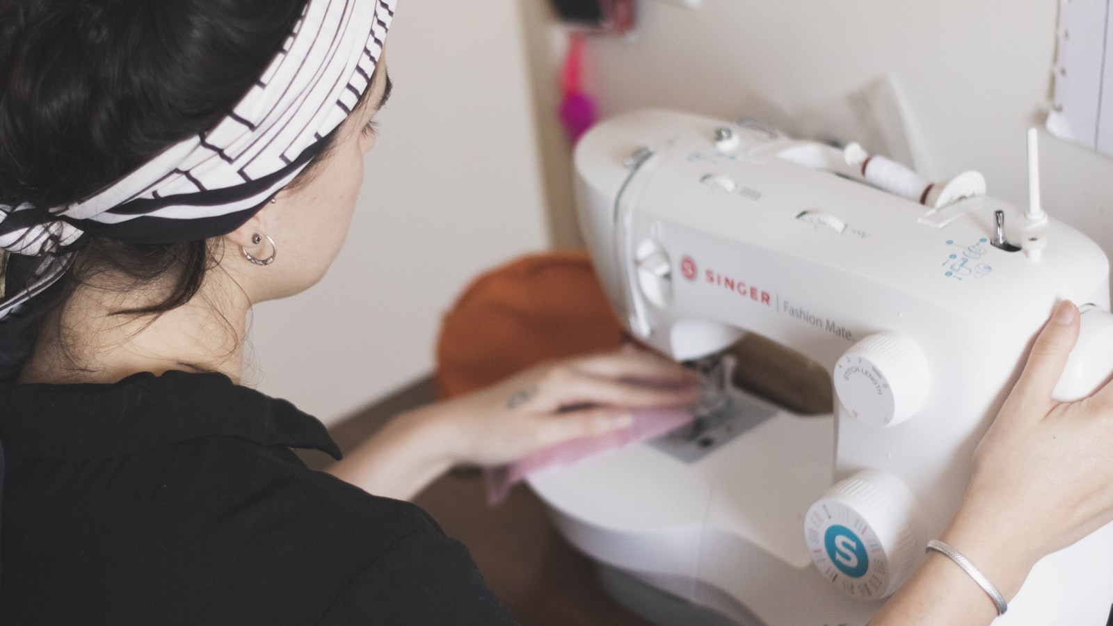 A person with a patterned headband guides pink fabric through a white sewing machine
