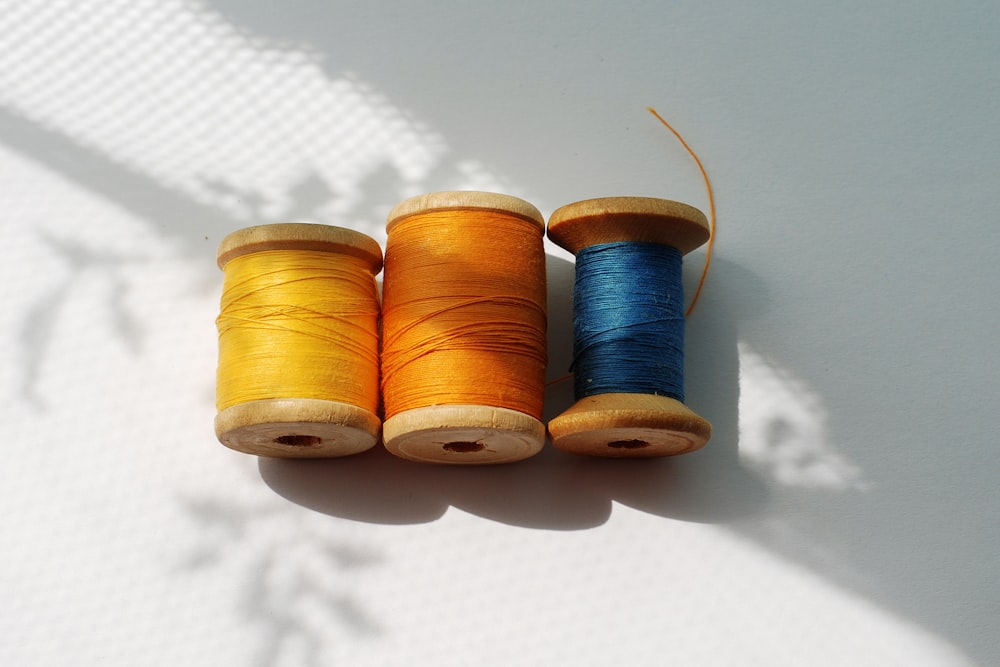 Three wooden spools of yellow, orange, and blue thread lie on a white surface