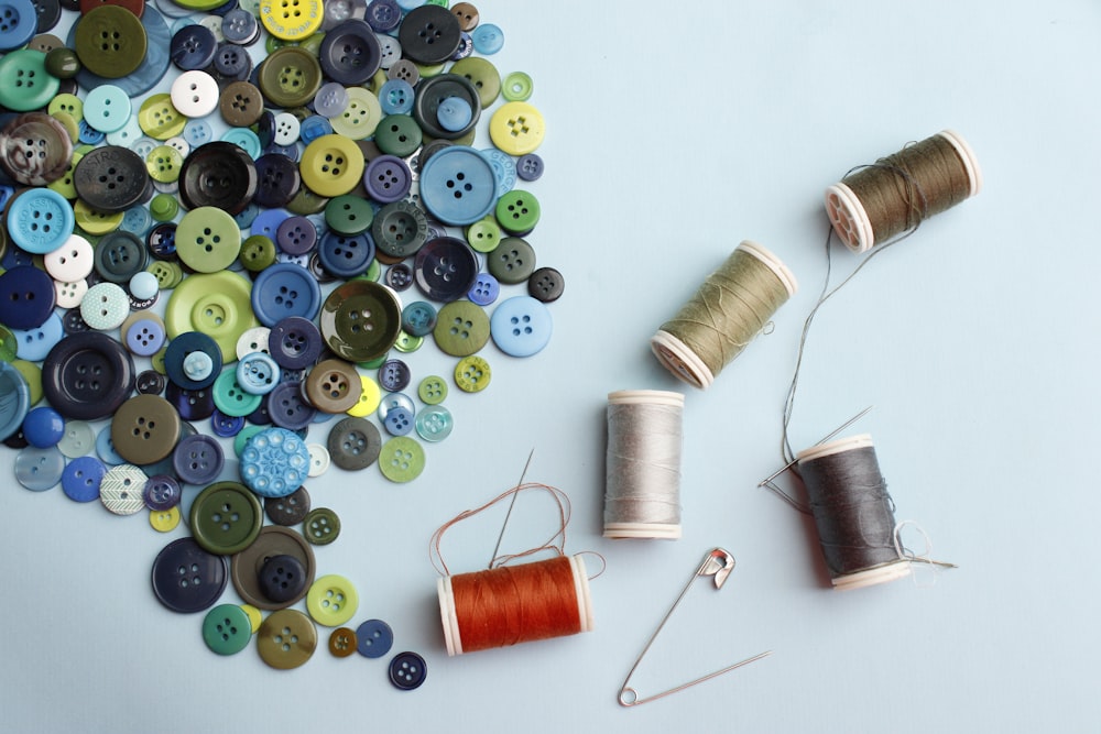 A pile of blue, green, and yellow buttons next to spools of thread and needles on a light blue surface
